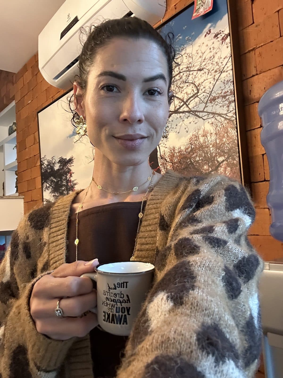 Camila Balieiro em casa, de casaco, segurando uma caneca.