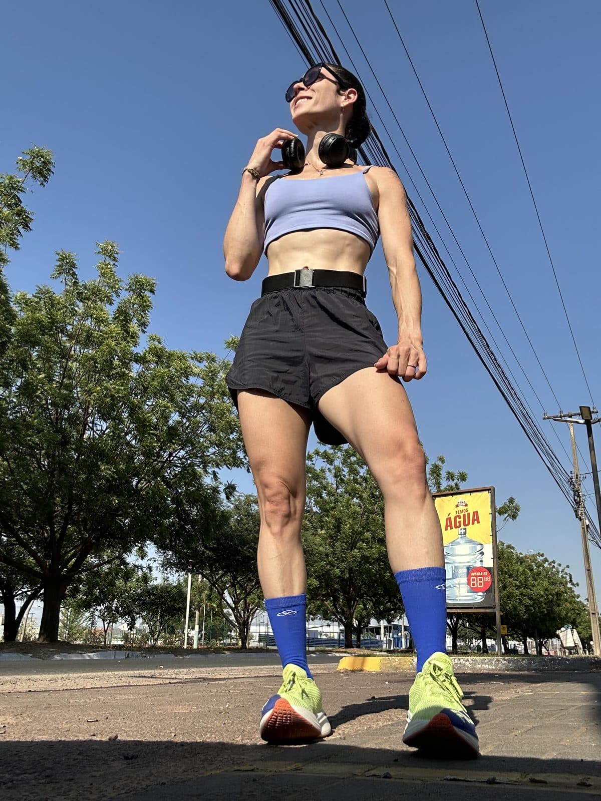 Camila Balieiro de roupa de treino e fones, em uma rua ensolarada, vista de baixo.