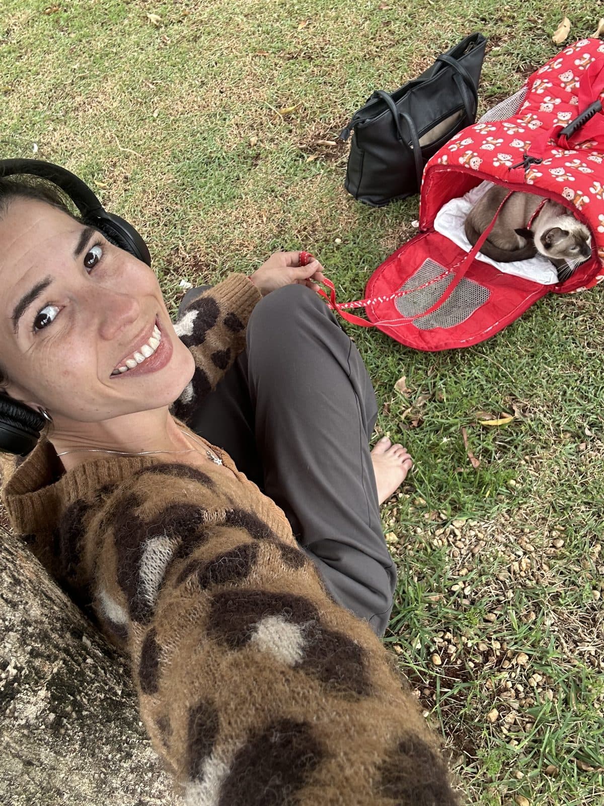 Camila Balieiro sitting on the grass with headphones, next to her cats' carrier, during the trip to Florianópolis.