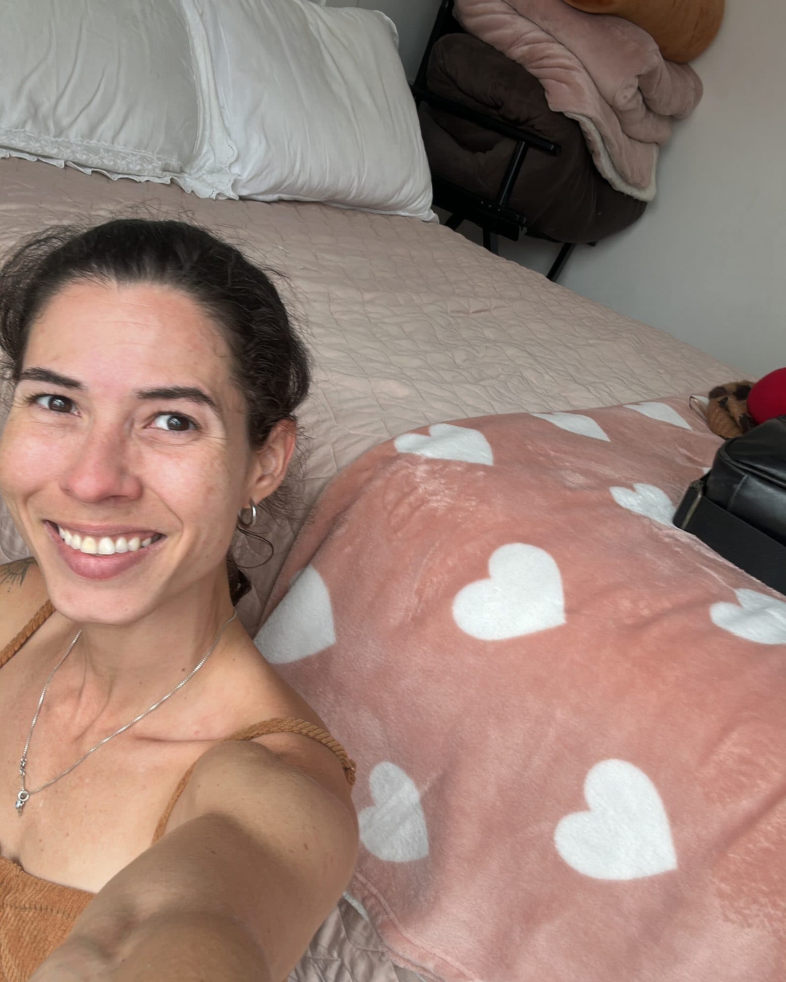 Camila sonríe recostada en la cama junto a una manta rosa con corazones.