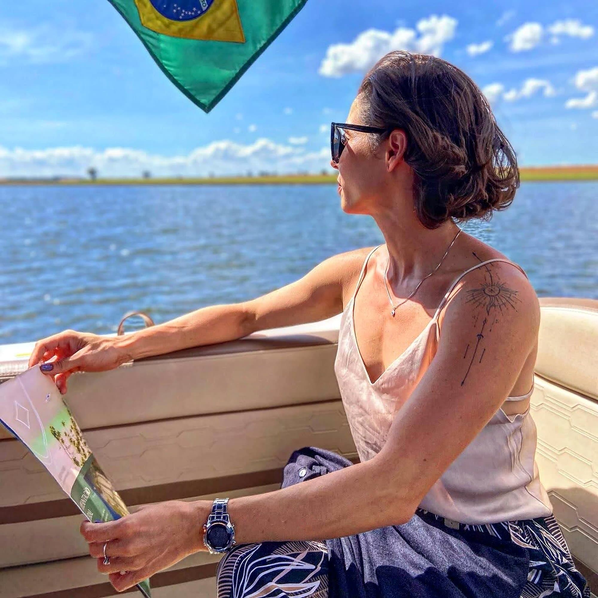 Camila con gafas de sol en un barco, mirando el agua, con la bandera de Brasil arriba.