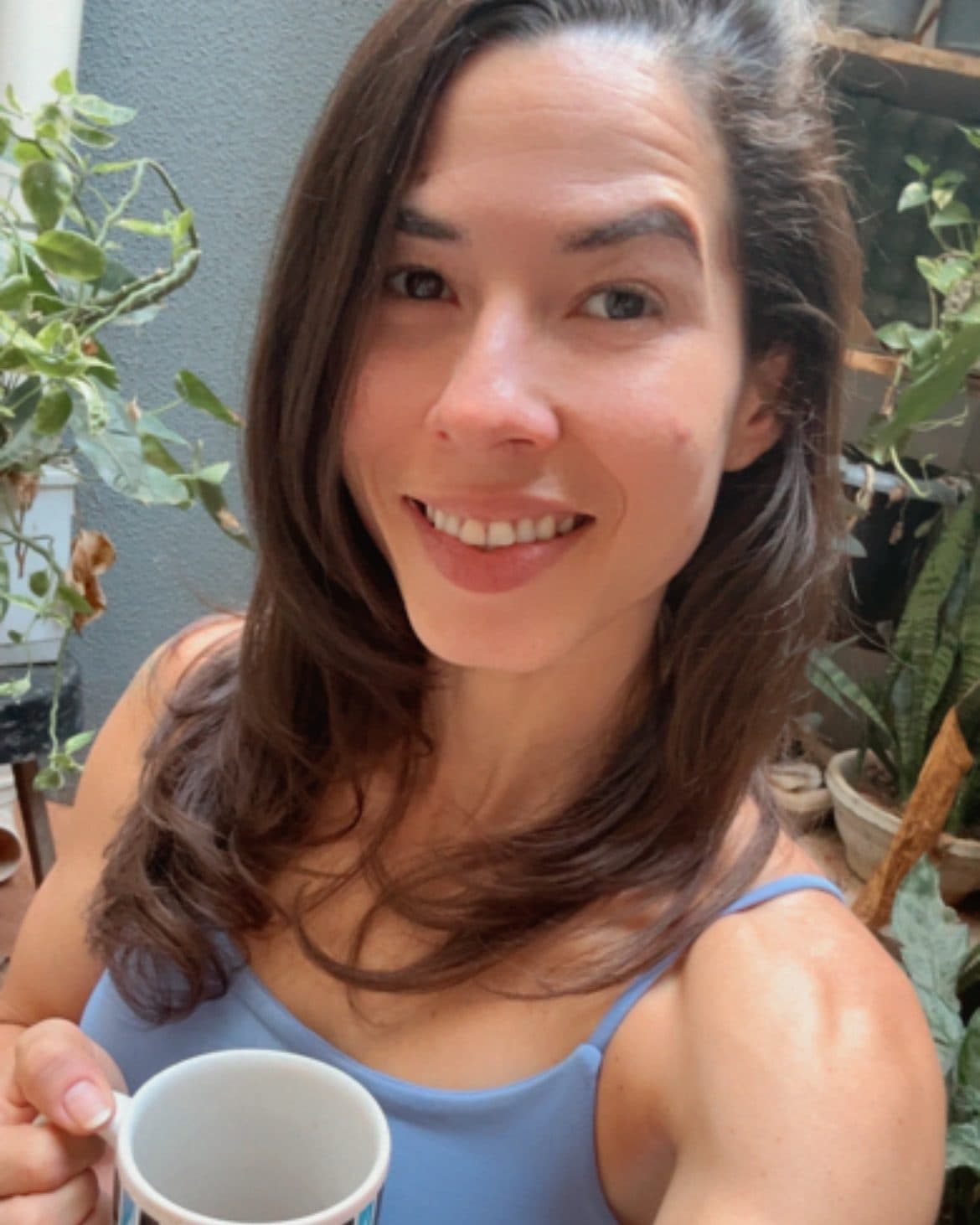 Camila Balieiro smiling with a coffee mug among plants.