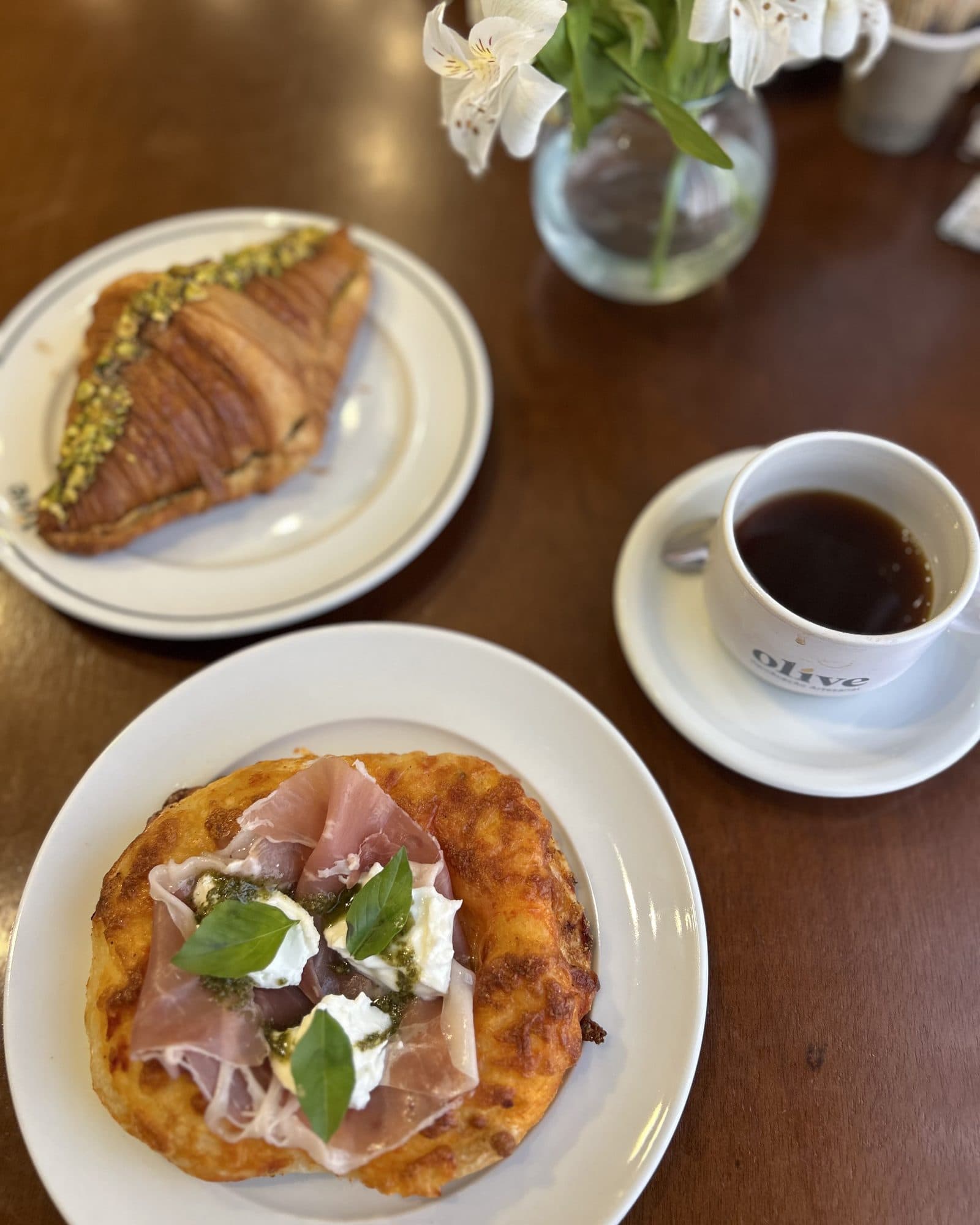 Mesa de café con un cruasán, una taza de café, una pizza con jamón y un florero.