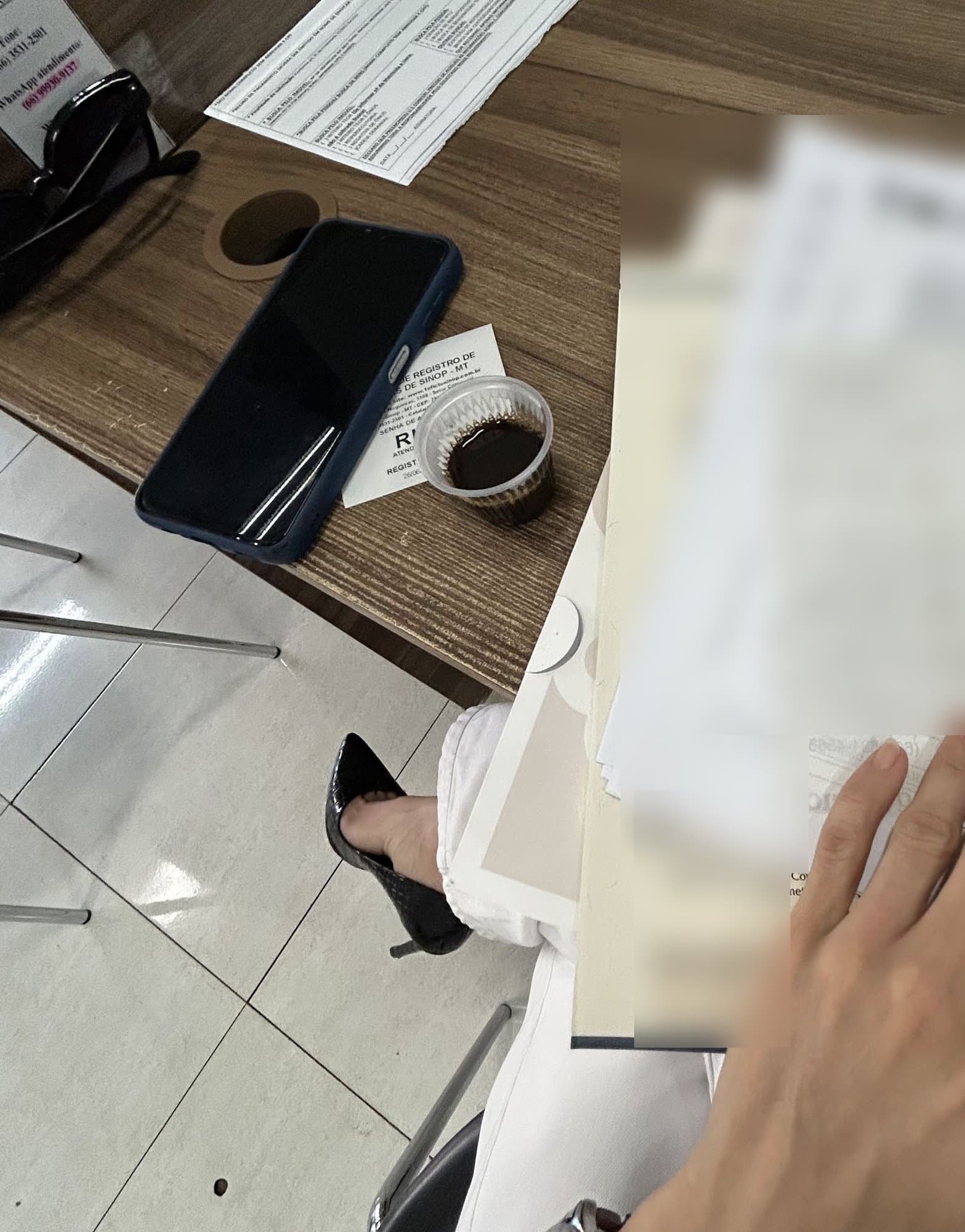 Vista desde arriba de una mesa de notaría: un móvil, gafas de sol, un vasito de café y un número de turno; a la derecha, una pila de documentos difuminados bajo la mano de una mujer con pantalón blanco y zapato de tacón negro.