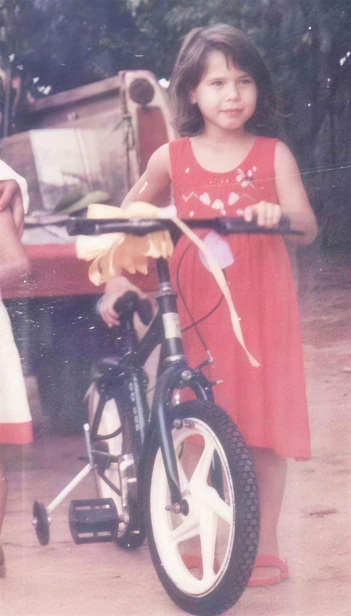 Camila de niña, con vestido rojo, junto a una bicicleta infantil con rueditas.