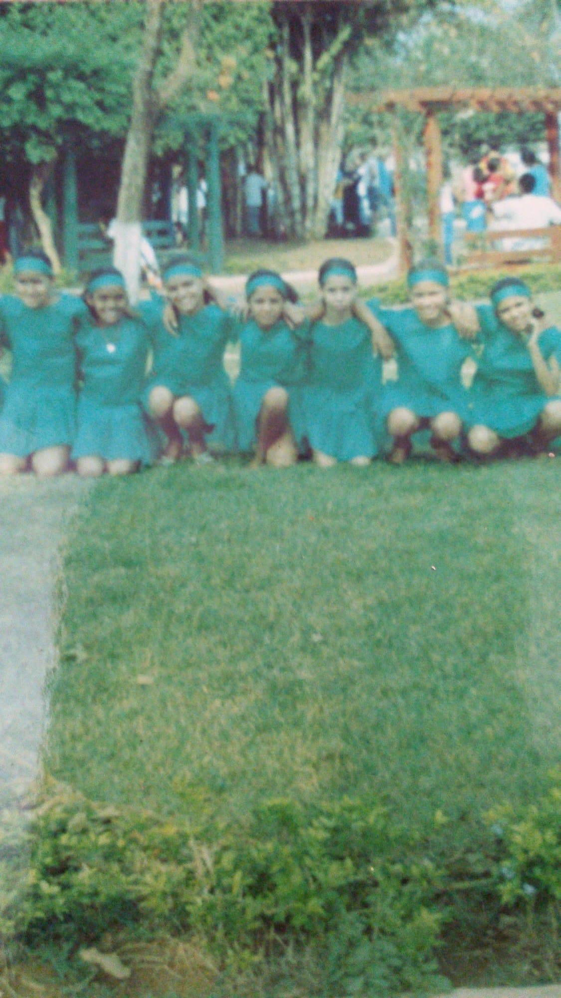 Niñas con uniforme verde y cinta en el pelo agachadas en fila sobre el césped, en la presentación del 7 de Septiembre.