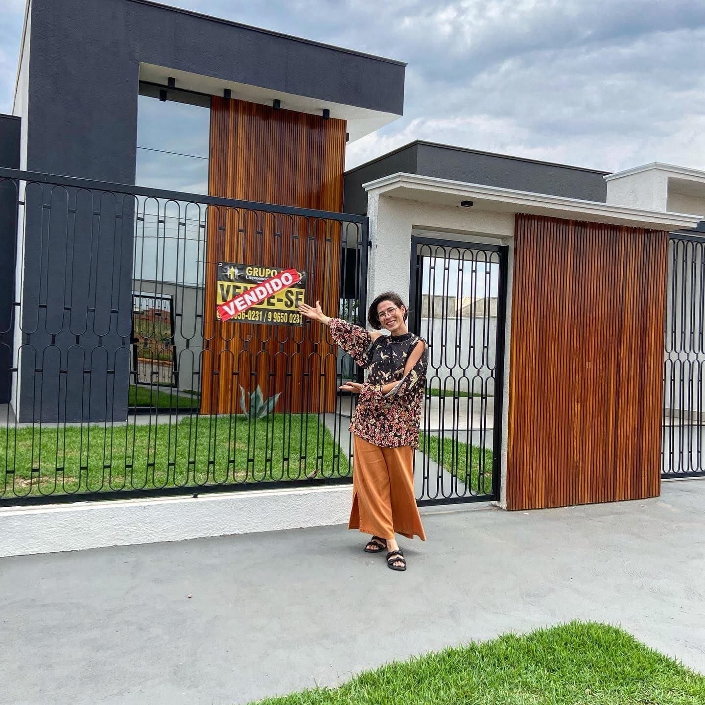 Camila, con blusa de flores y pantalón naranja, sonríe y señala un cartel de 'Se vende' con la franja 'Vendido' en la reja de una casa moderna con listones de madera.
