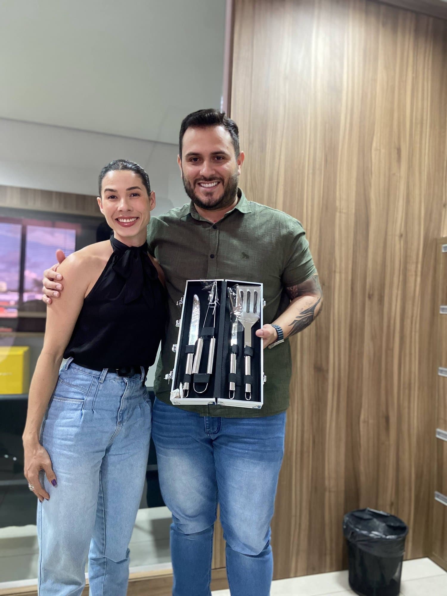 Camila sonríe abrazada a un hombre que sostiene un set de parrilla en su estuche, en una oficina con panel de madera.