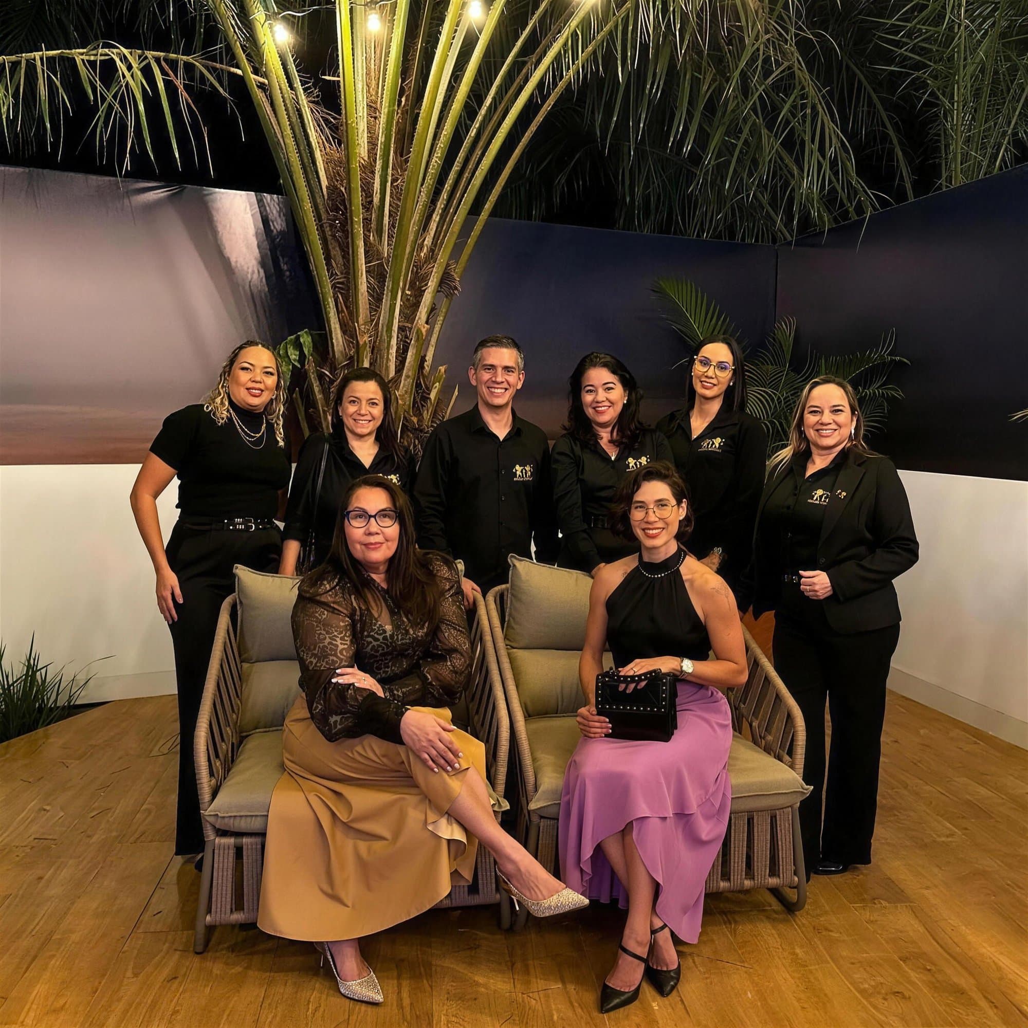 Equipo de nueve personas, casi todas de negro, posando alrededor de dos sillones bajo una palmera, de noche; Camila sentada con falda lila.