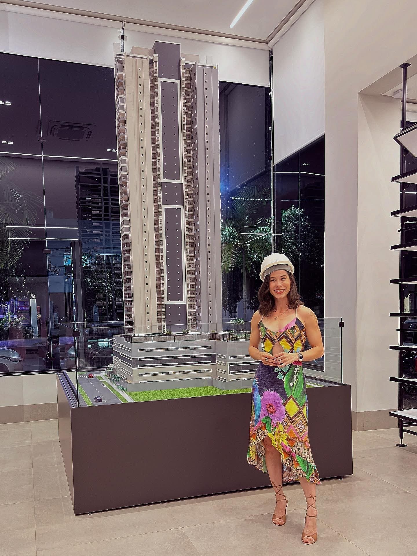 Camila con vestido estampado colorido y gorra blanca junto a la maqueta de una torre residencial alta, en una sala de ventas.