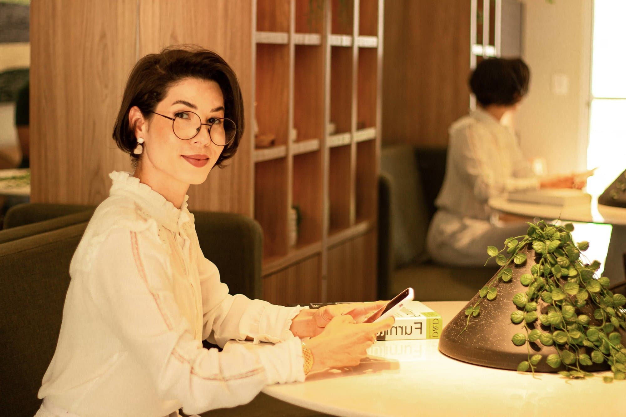 Camila de pelo corto, gafas y blusa blanca, sentada a una mesa con el móvil en la mano, sonriendo a la cámara; al fondo trabaja otra mujer.