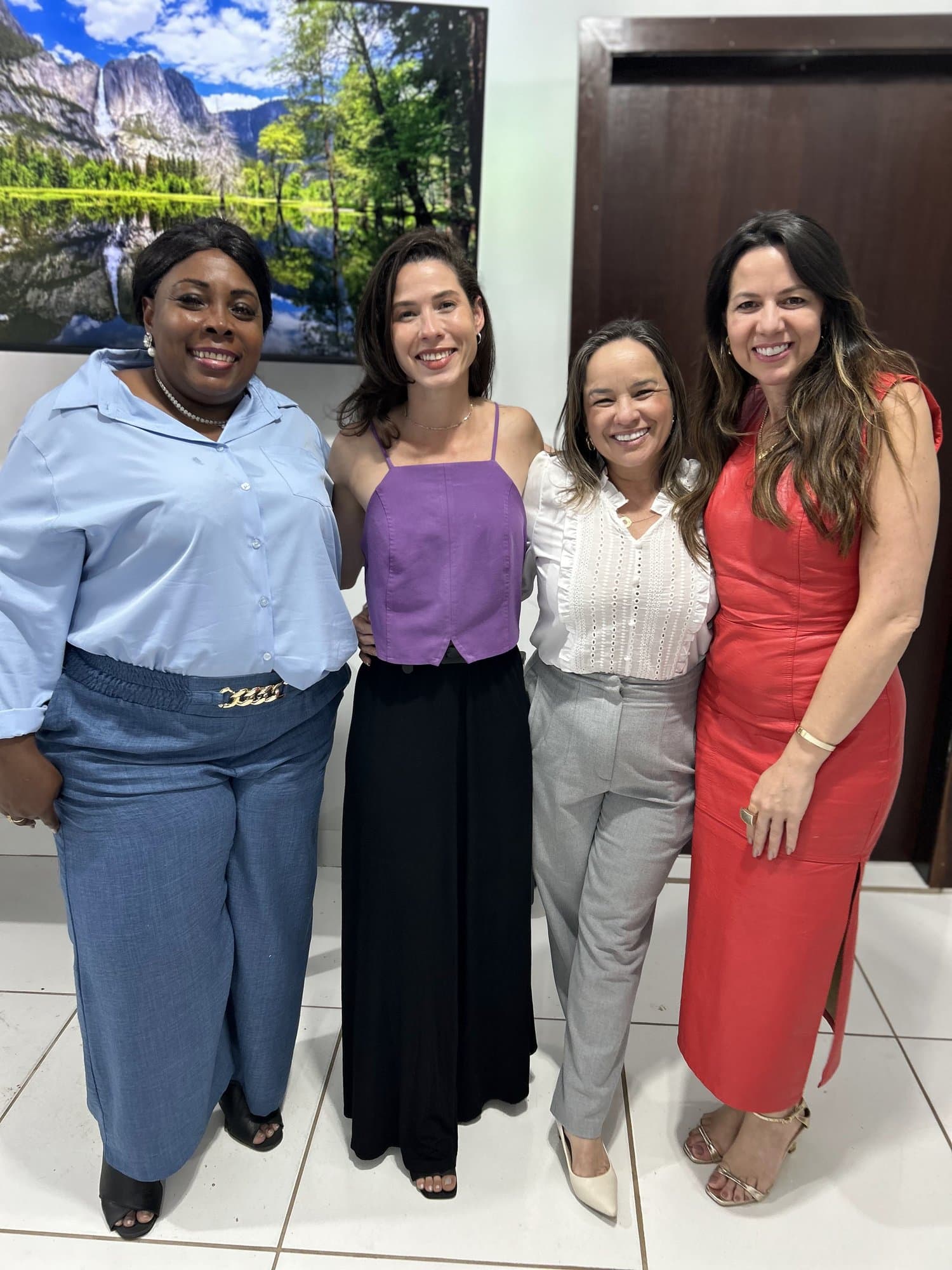 Cuatro mujeres sonriendo una al lado de otra frente a un cuadro de montañas y lago; Camila lleva blusa lila y pantalón negro.