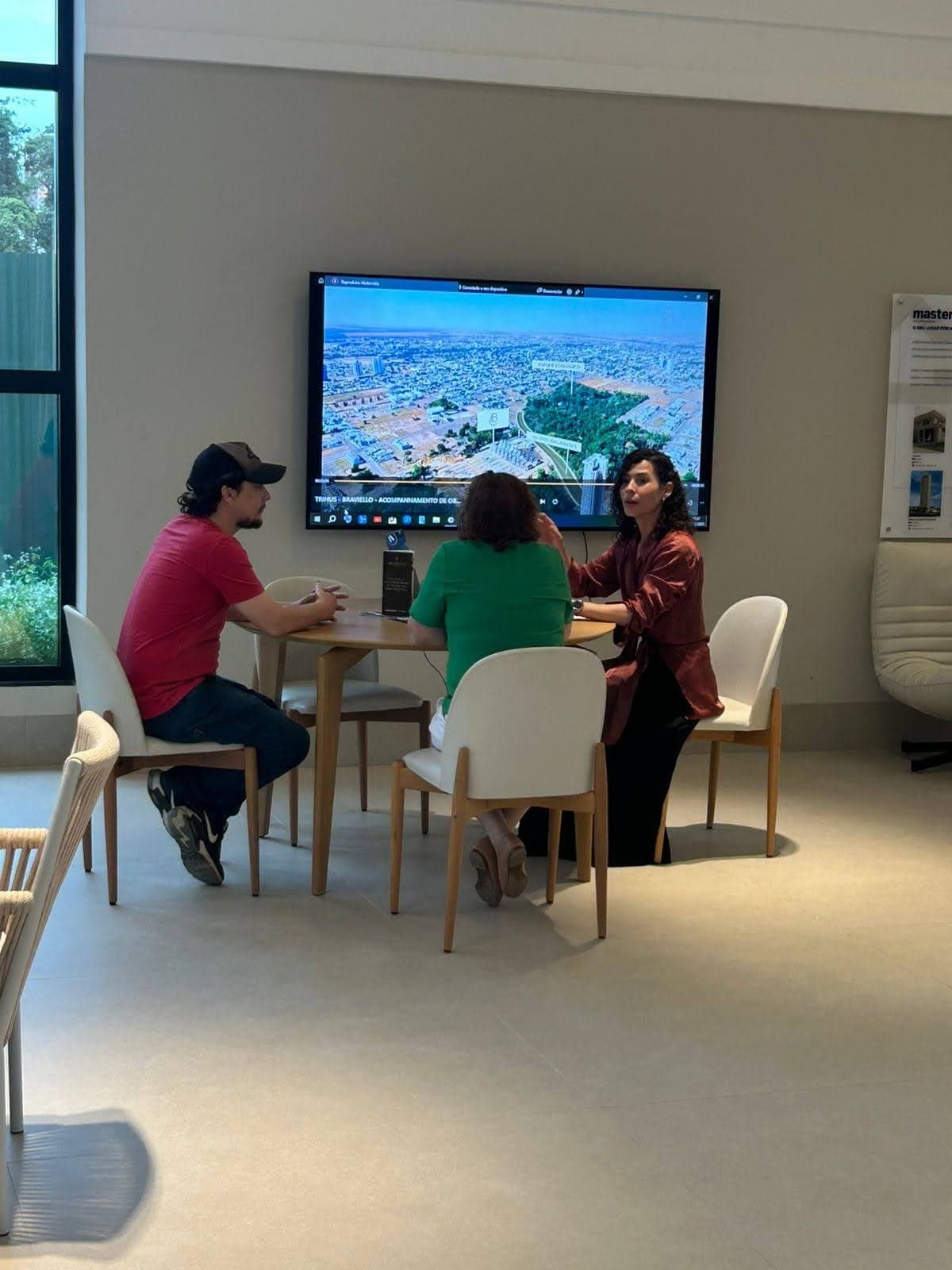 Camila conversa con dos clientes en una mesa redonda de una sala de ventas, frente a una TV con una vista aérea de la ciudad.
