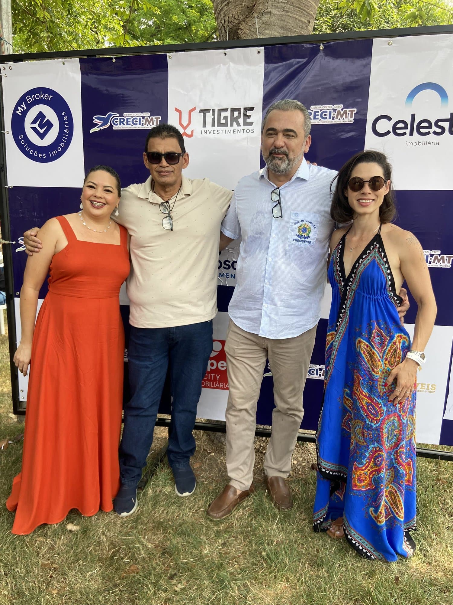 Camila con gafas de sol y vestido azul estampado posa con una mujer de naranja y dos hombres frente a un panel con logos del CRECI-MT y de inmobiliarias, al aire libre.