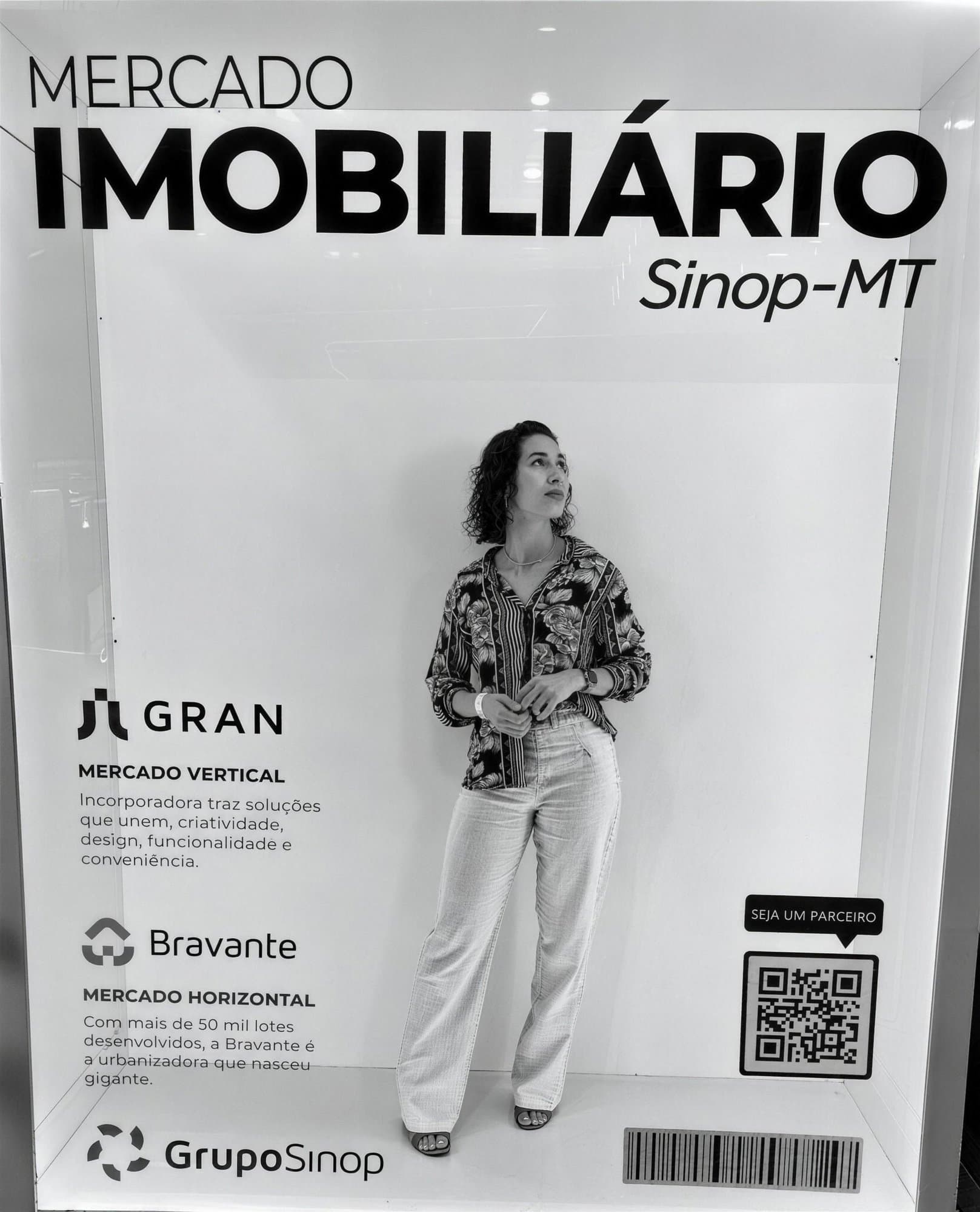 Foto en blanco y negro: Camila de pie dentro de un panel expositor titulado "Mercado Imobiliário Sinop-MT" con logos de Gran, Bravante y Grupo Sinop.