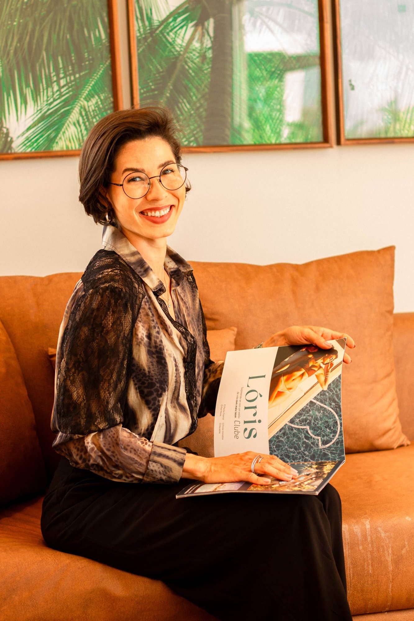 Camila con gafas y blusa estampada, sonriendo en un sofá naranja, sostiene abierto el folleto del emprendimiento Lóris.