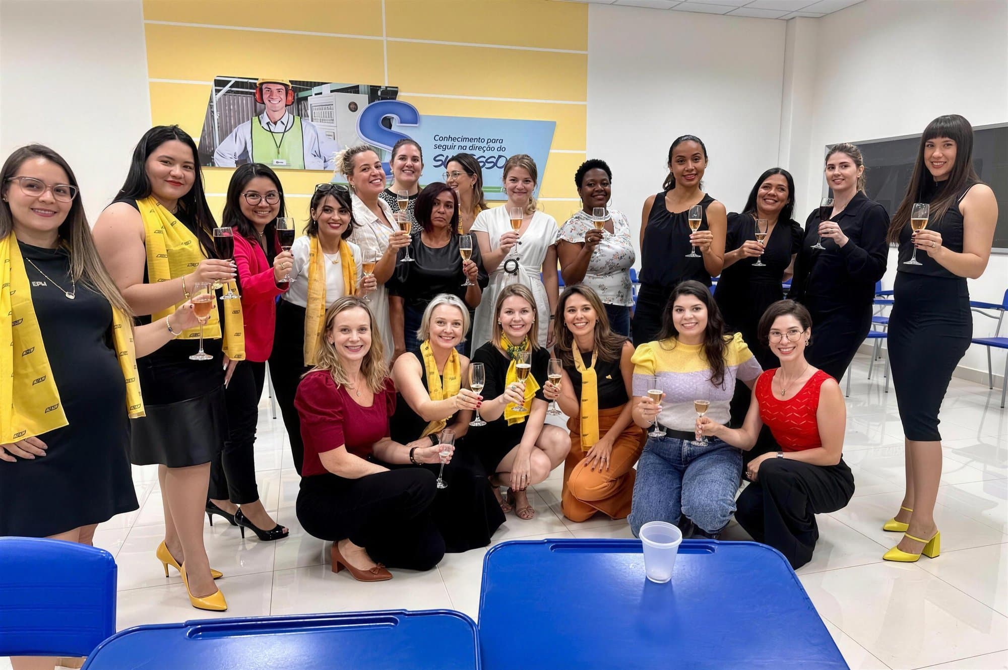 Grupo de unas veinte mujeres, varias con pañuelos amarillos, brindando con copas en una sala de formación.