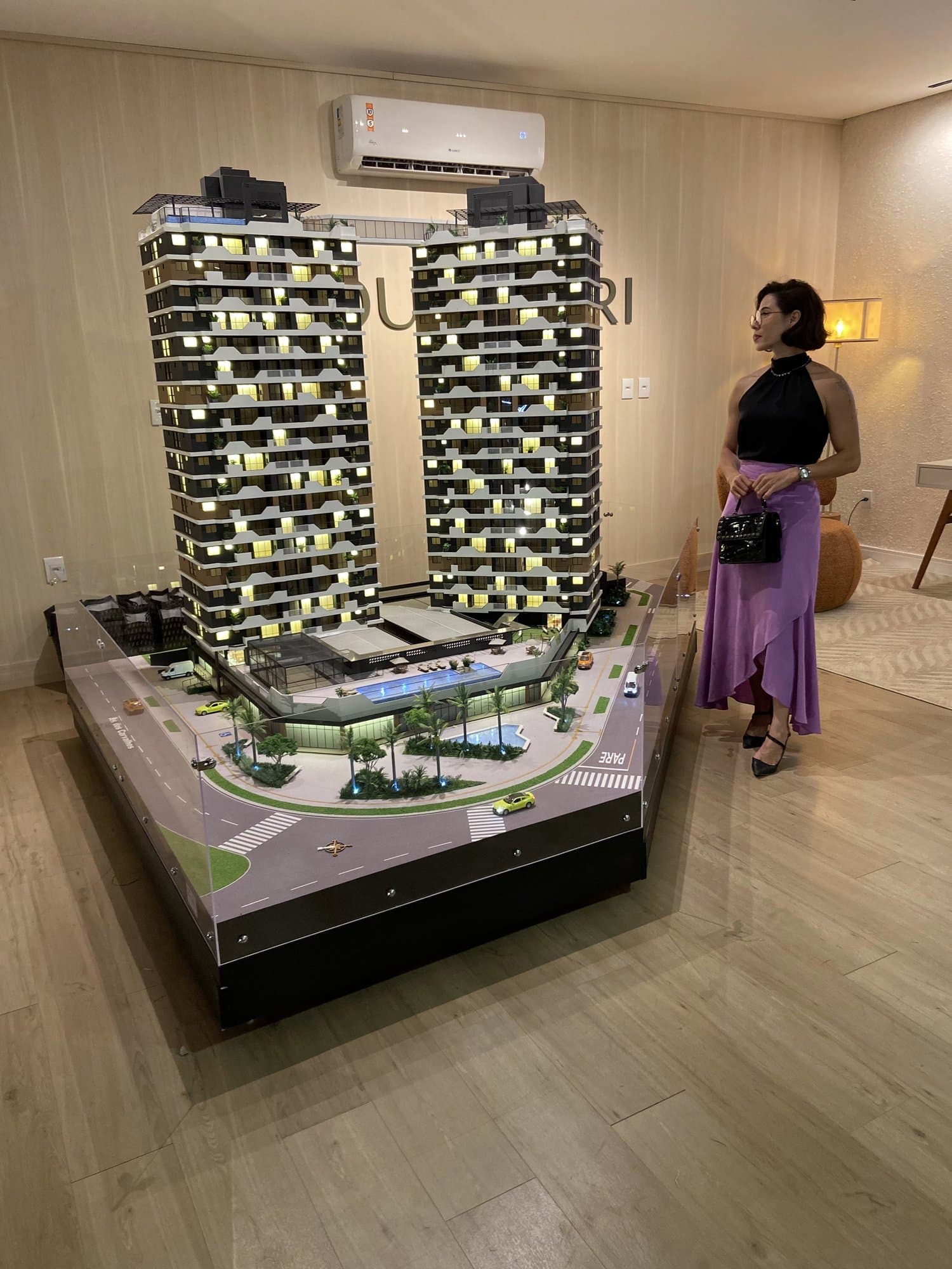 Maqueta iluminada de dos torres residenciales con área de ocio; a la derecha, Camila, con blusa negra y falda lila, la observa.