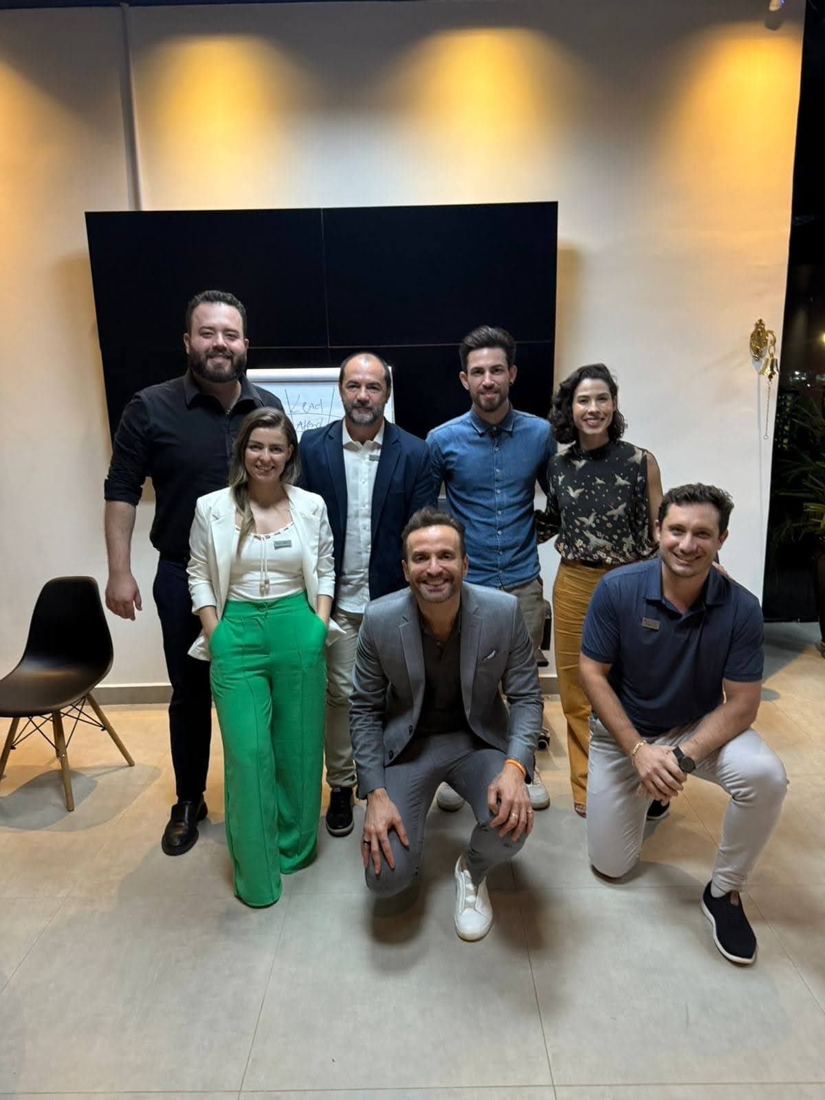 Grupo de ocho personas posando en una sala con pared oscura, algunas agachadas delante; Camila con blusa estampada a la derecha.