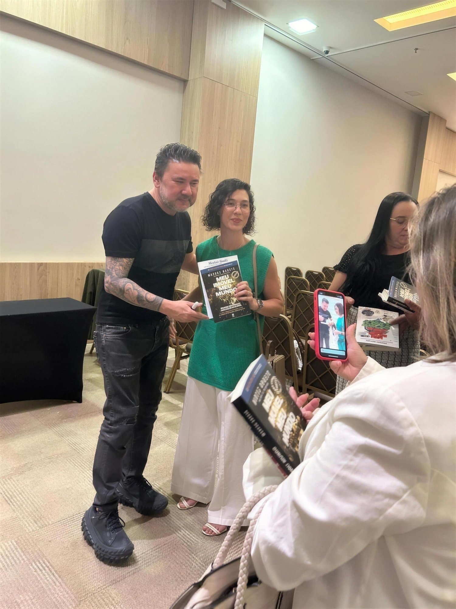 Camila, con blusa verde, sostiene un libro junto a un hombre tatuado en un evento; delante, una mujer de espaldas toma una foto con el móvil.