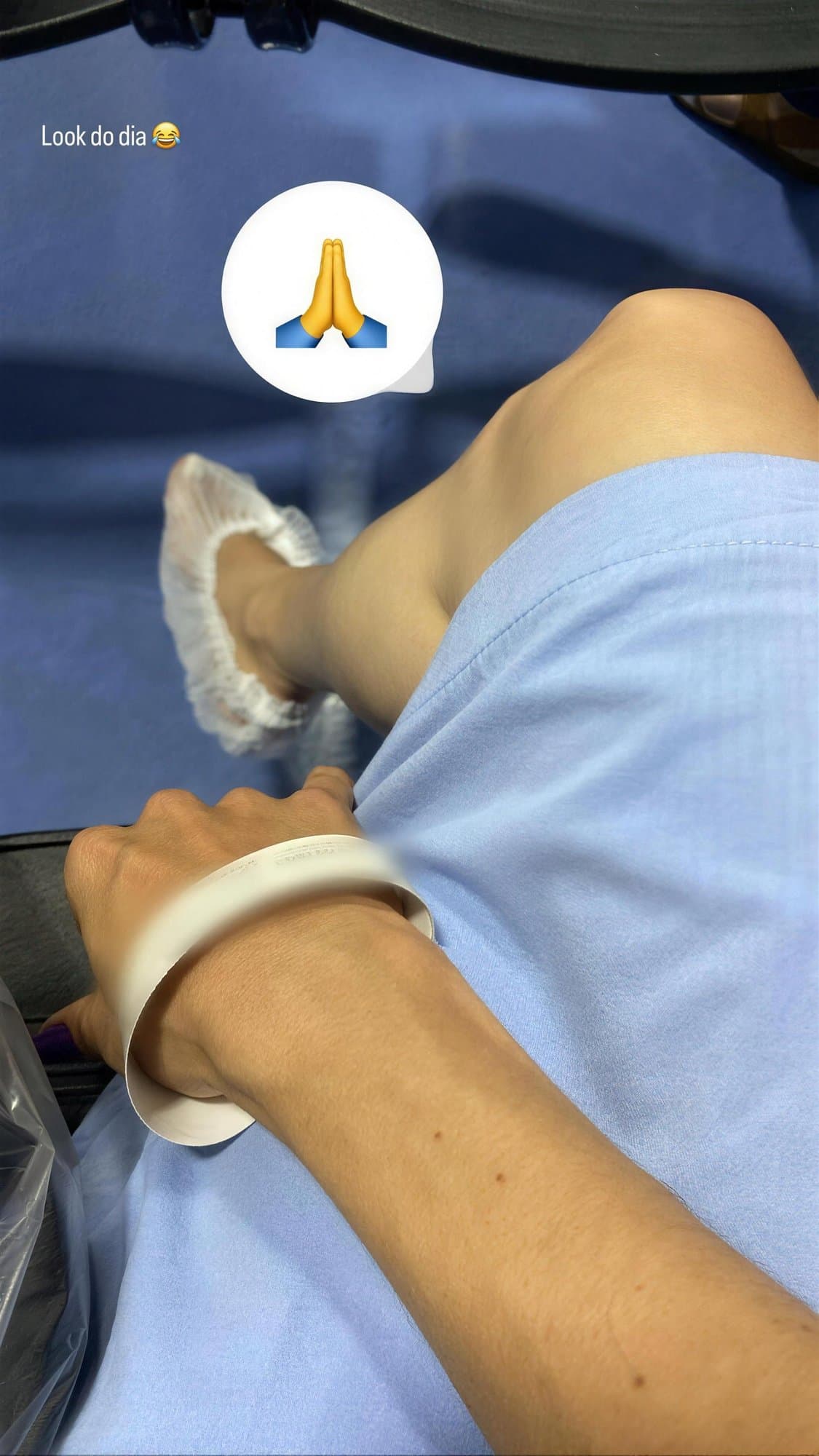 Vista desde arriba de un brazo con pulsera hospitalaria blanca sobre una bata azul, y un pie con cubrezapato desechable.
