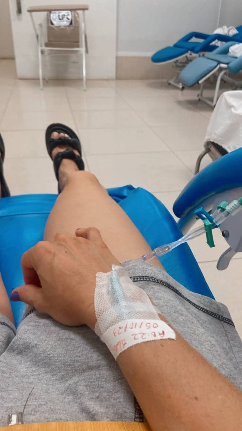 Una mano con vía intravenosa y esparadrapo, conectada a un equipo, apoyada en la pierna, en una sala de infusión con sillones azules.