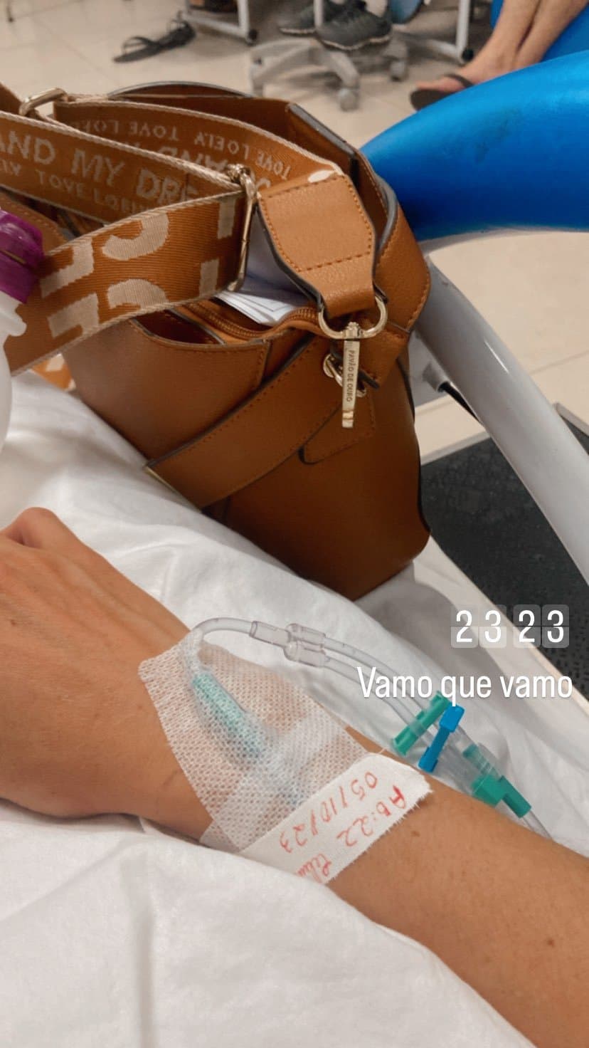 Una mano con vía intravenosa sobre una sábana blanca, junto a un bolso color caramelo, en una sala de infusión.