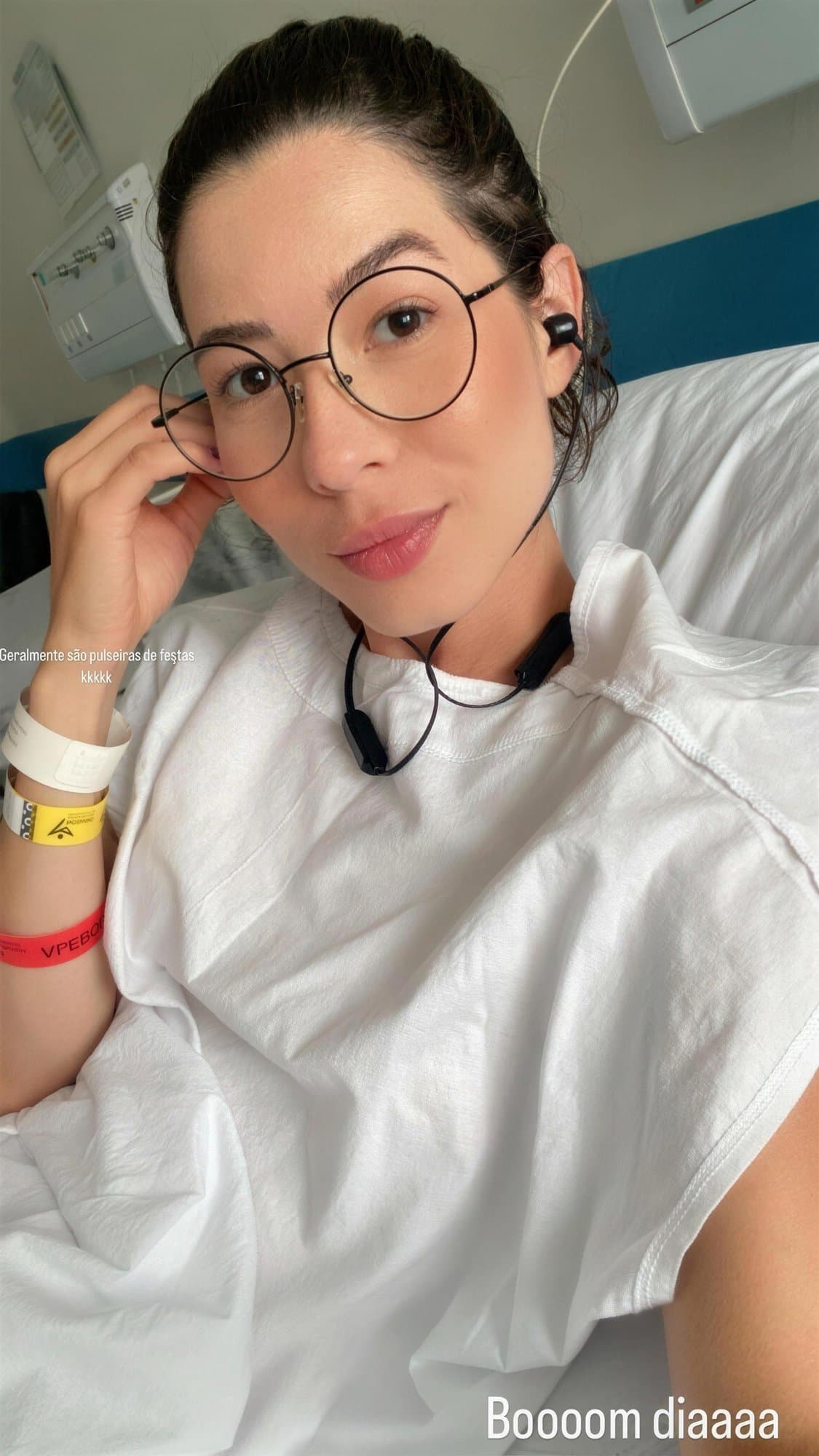 Selfie de Camila con gafas redondas y bata hospitalaria blanca, acostada en una cama de hospital, con pulseras hospitalarias en la muñeca.