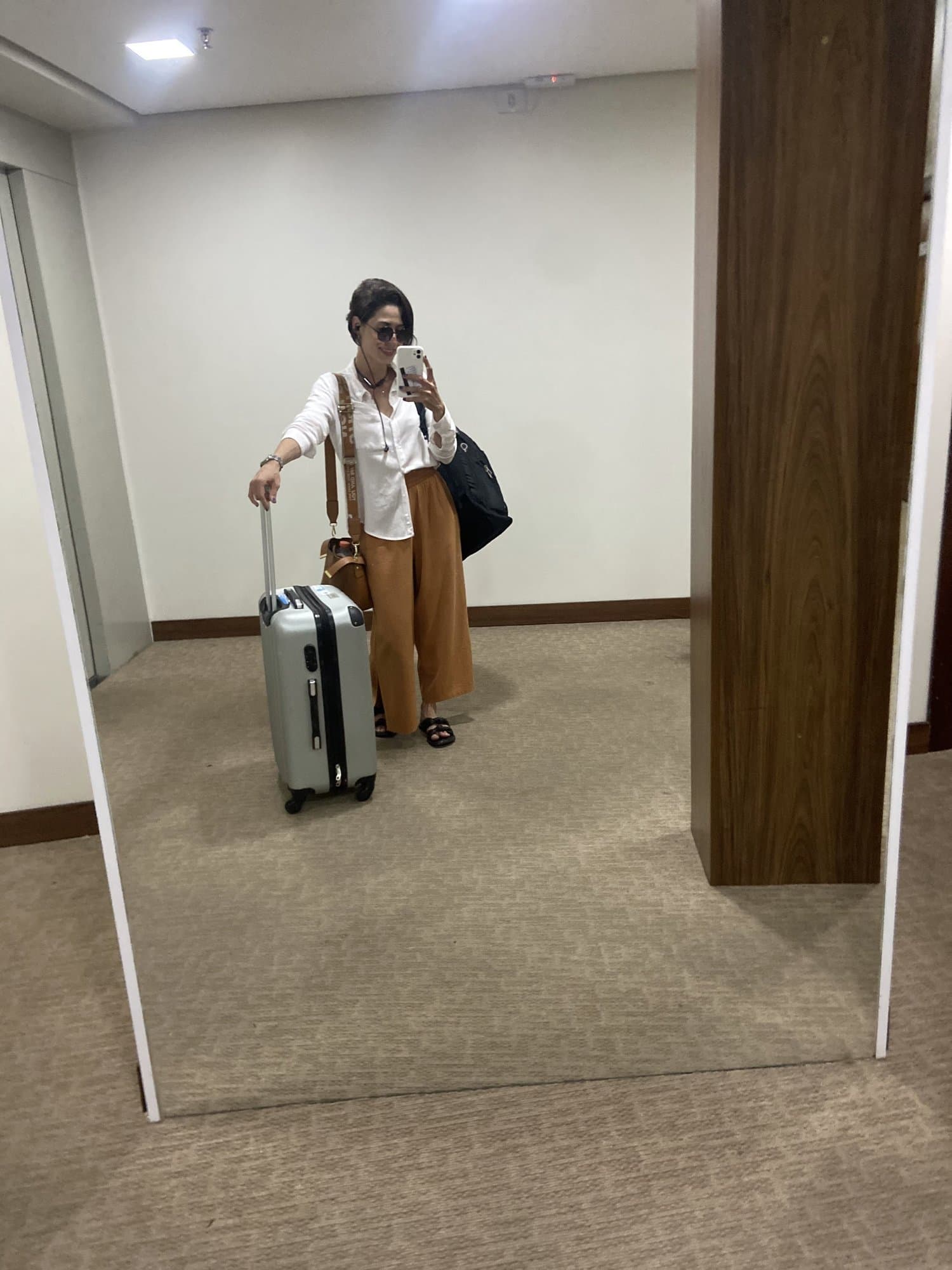 Selfie en el espejo: Camila con camisa blanca, pantalón caramelo y gafas de sol en la cabeza, con maleta de ruedas y bolsos, en un pasillo alfombrado.