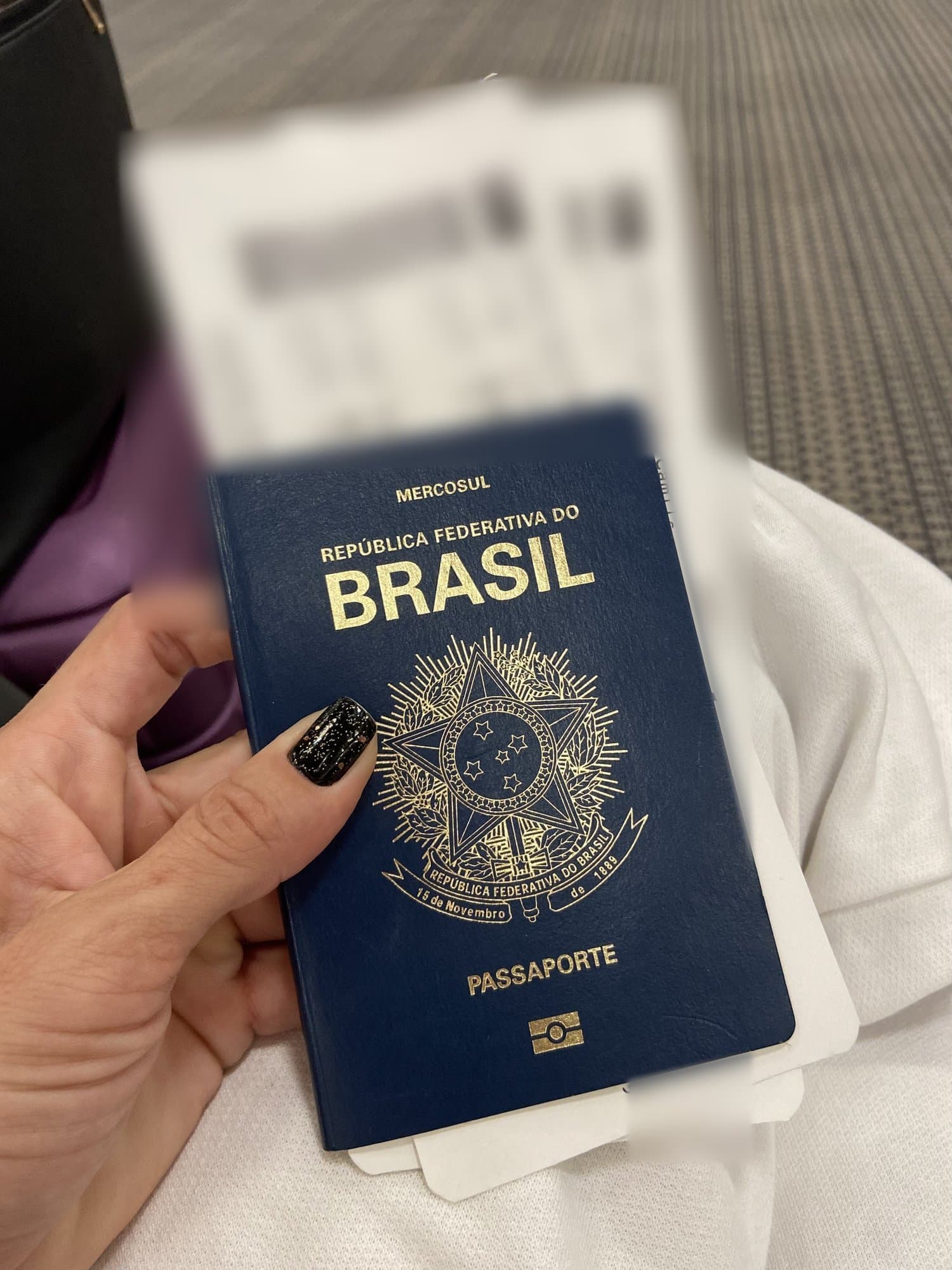 Una mano con uñas negras sosteniendo un pasaporte brasileño y tarjetas de embarque.