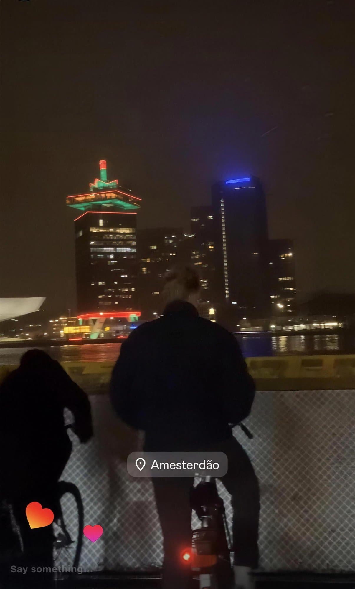 Dos personas en bicicleta, de espaldas, mirando edificios iluminados de noche al otro lado del agua en Ámsterdam.