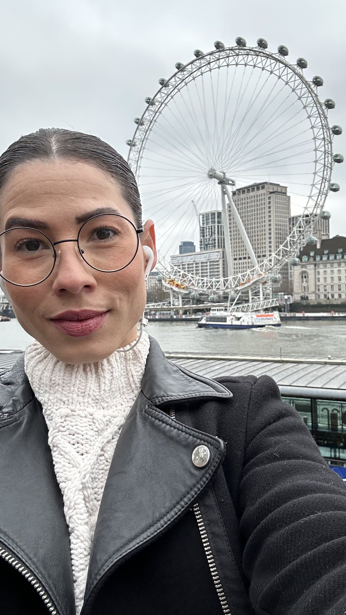 Camila con gafas y suéter blanco en un selfie junto al Támesis, con el London Eye detrás.