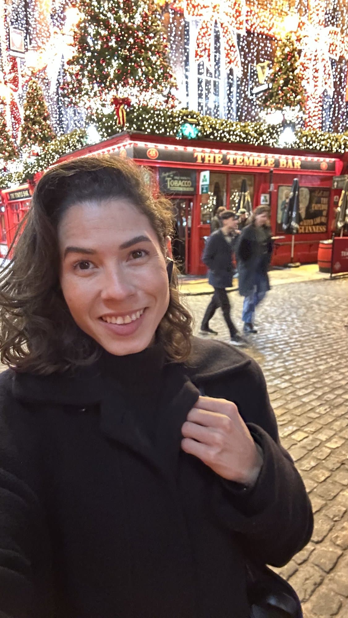 Camila sonriendo con abrigo negro frente al pub The Temple Bar de Dublín, decorado con luces de Navidad.