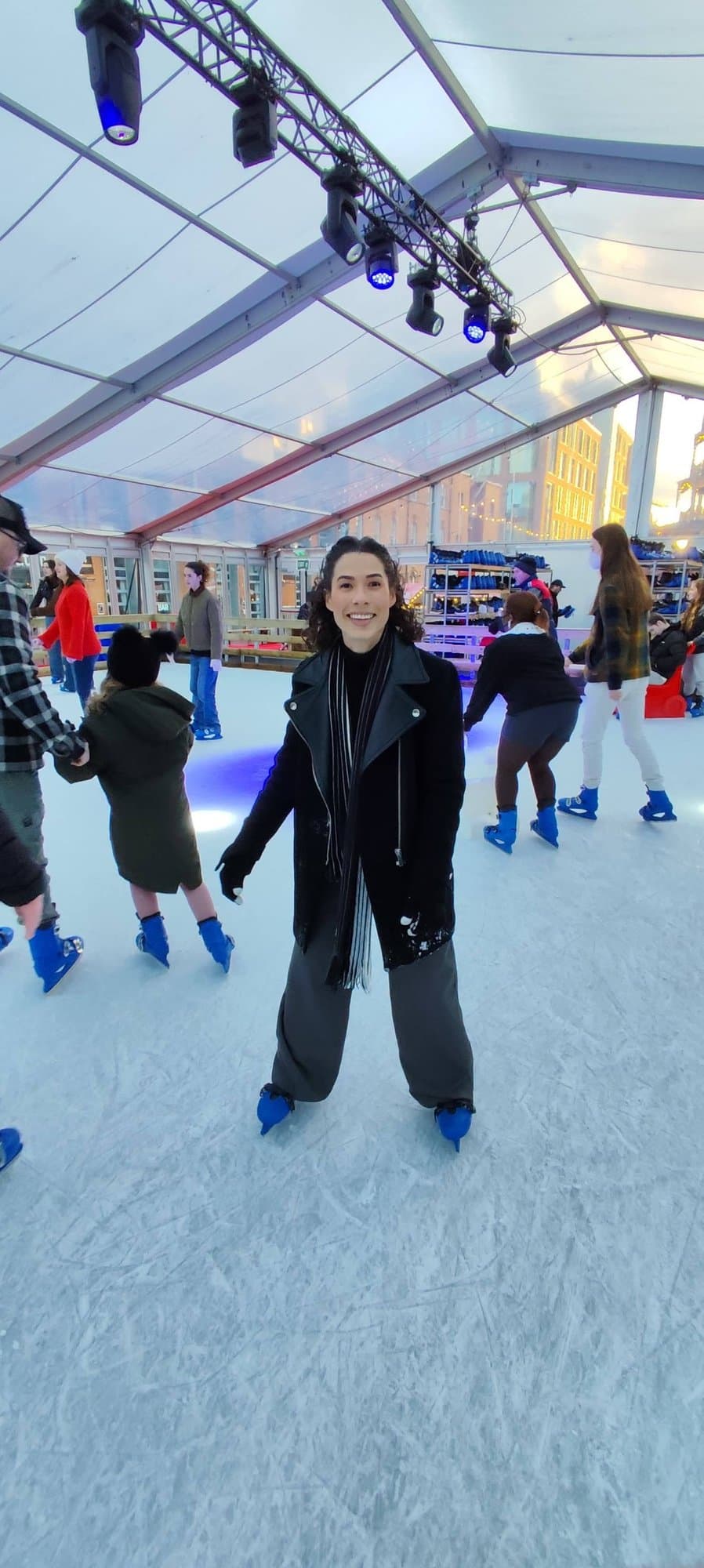 Camila con abrigo negro patinando en una pista de hielo cubierta, entre otros patinadores.