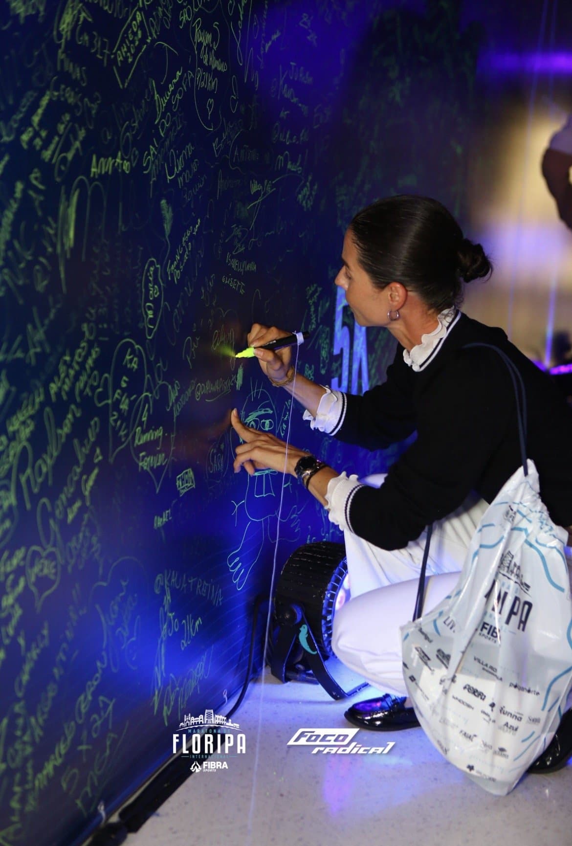 Camila agachada escribiendo con un rotulador neón en un mural lleno de firmas, bajo luz azul, con la bolsa del kit de la carrera.