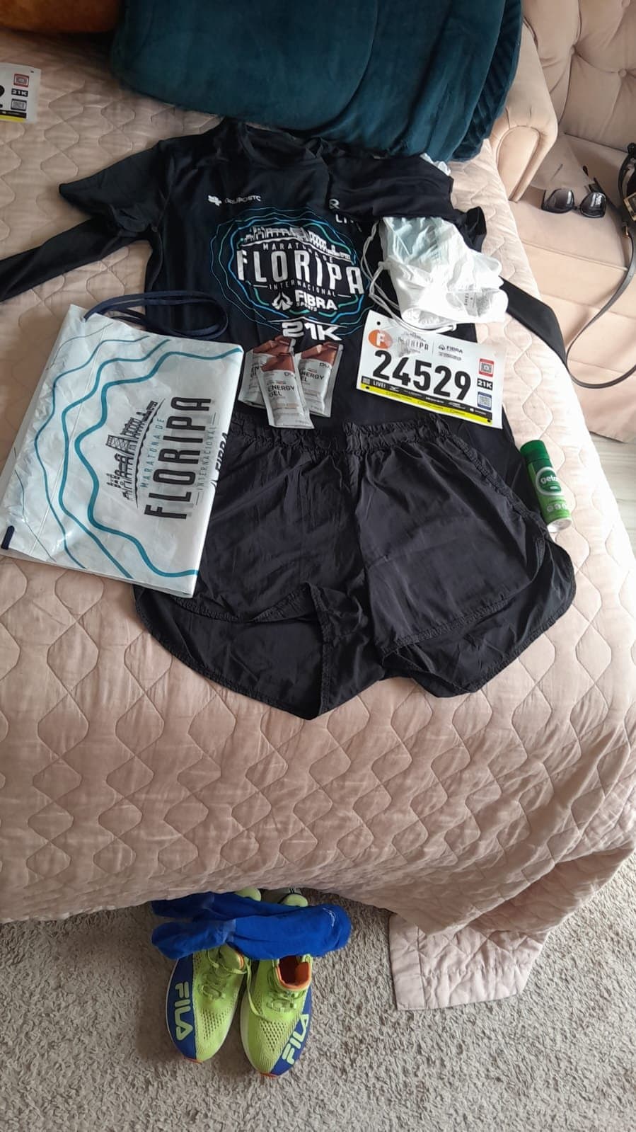 Kit de la media maratón sobre la cama: camiseta Floripa 21K, short, dorsal 24529, geles y zapatillas verdes en el suelo.