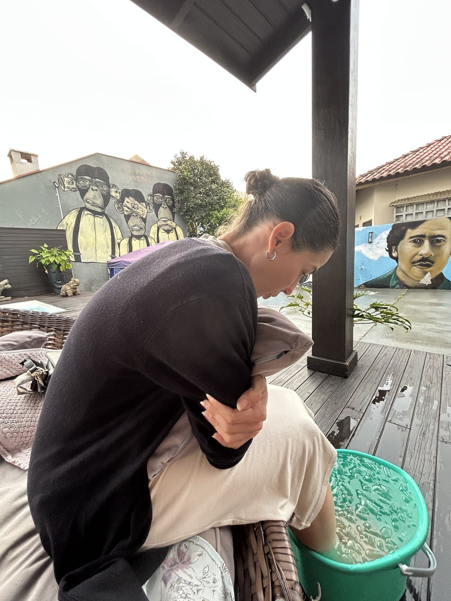 Camila sentada en un porche con los pies en un balde de agua con hielo, abrazando un cojín; murales de grafiti al fondo.