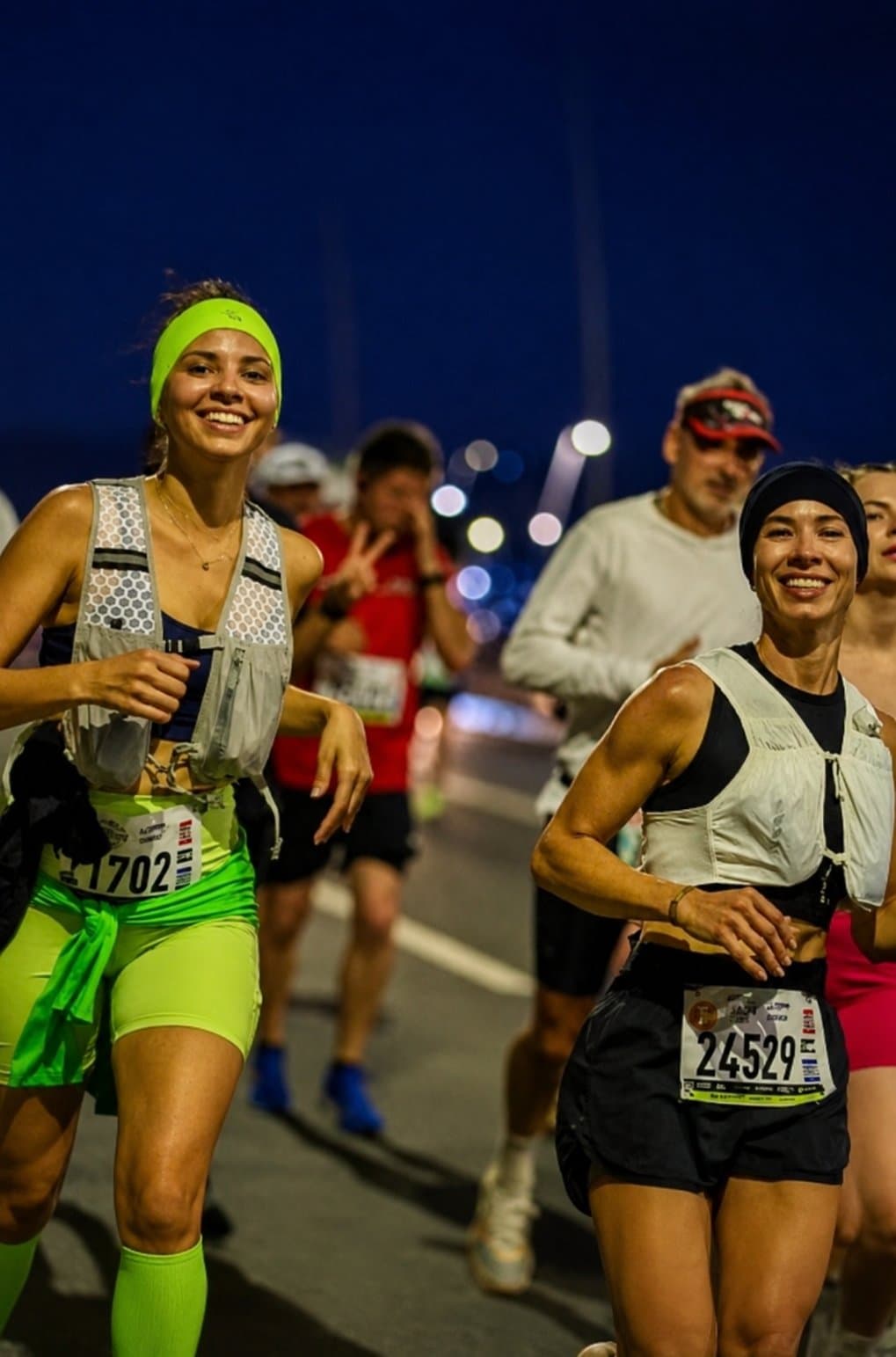 Camila sonriendo en la salida nocturna de la carrera, con chaleco blanco y dorsal 24529, junto a otros corredores.