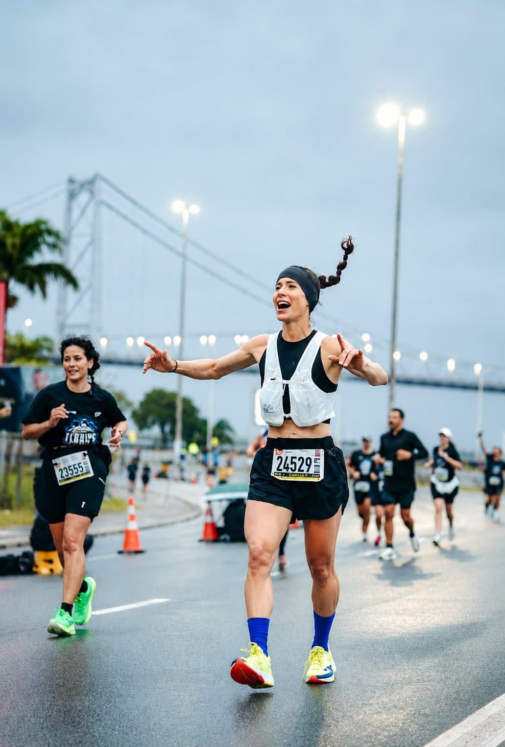 Camila corriendo con los brazos abiertos y sonriendo en una avenida mojada, con el puente Hercílio Luz al fondo.