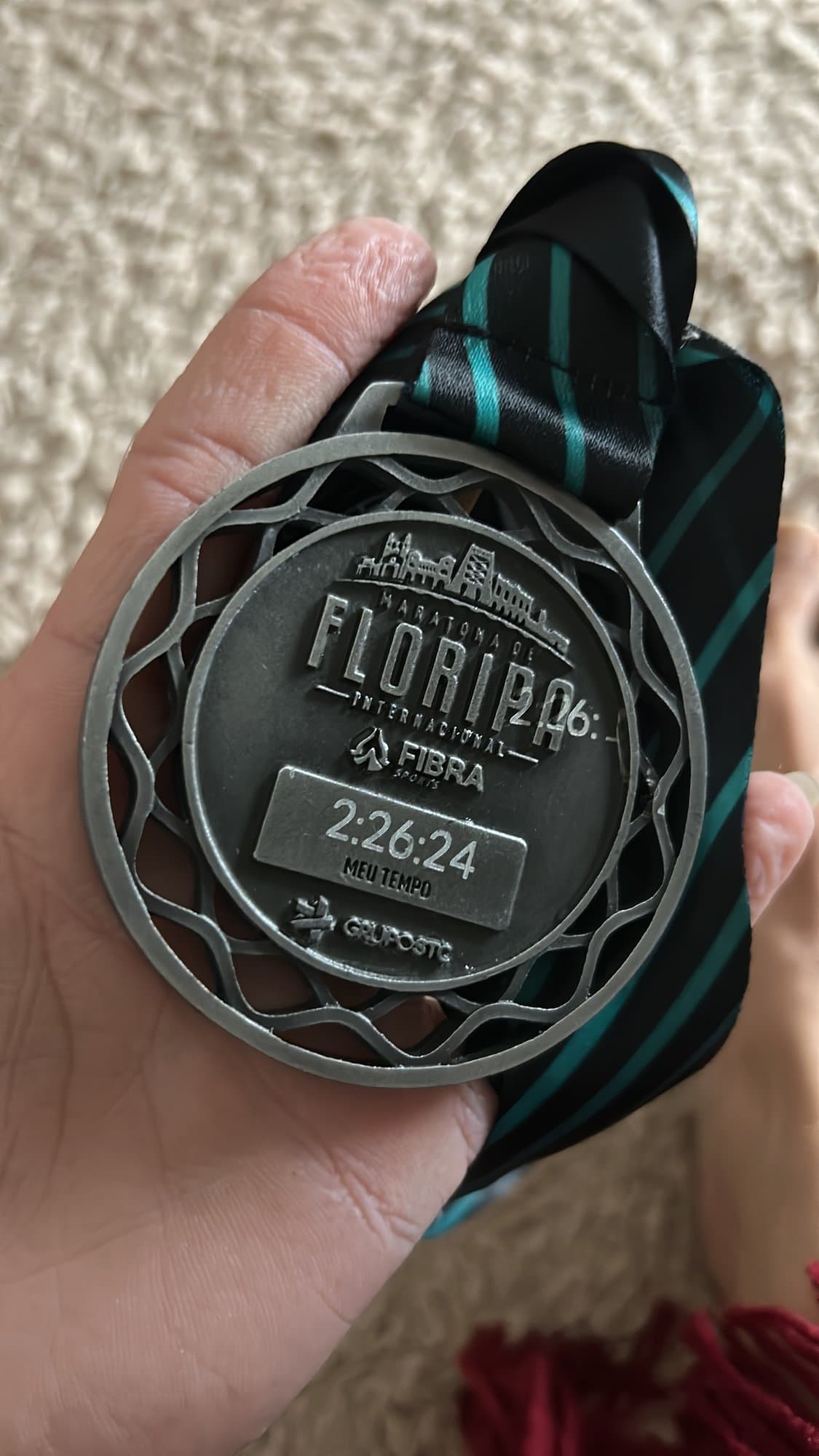 Una mano sosteniendo la medalla de la Maratón Internacional de Floripa con el tiempo 2:26:24 grabado.